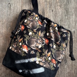 May include: Black drawstring bag with a woodland rabbit print. The bag has a black base with white spray paint-like accents. The main body of the bag is patterned with brown rabbits, red mushrooms, and green foliage.