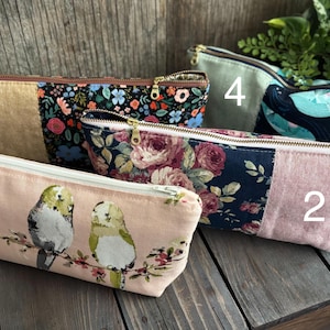 Peut inclure: Collection de cinq pochettes à fermeture éclair en tissu avec divers motifs. Les motifs comprennent des imprimés floraux, d'oiseaux et de baleines. Chaque pochette a une palette de couleurs différente, dont le rose, le bleu et le vert. Les pochettes sont disposées sur une surface en bois.