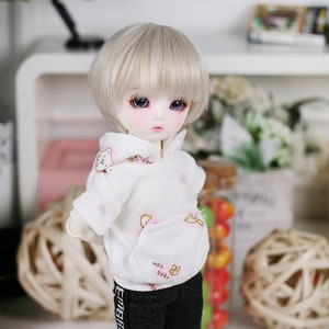 May include: A white doll with blonde hair wearing a white hoodie with a pink and white design and black pants with white lettering. The doll is wearing pink and white sneakers.