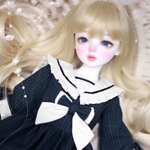 May include: A ball-jointed doll with long blonde hair and blue eyes, wearing a dark navy blue dress with white pinstripes, a white sailor-style collar with ruffles, a white bow at the chest, and white lace trim at the cuffs and hem.