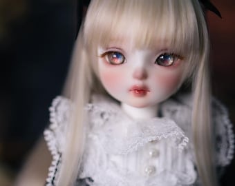 May include: A ball-jointed doll with pale skin, long straight platinum blonde hair, and large iridescent eyes. The doll wears a black dress featuring a high white lace collar with small pearl-like buttons and white lace trim on the sleeves.