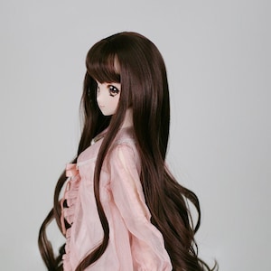 May include: A long, brown wig with bangs styled in loose waves. The wig is worn by a doll with a pink dress and white underskirt. The doll has brown eyes and a light complexion.