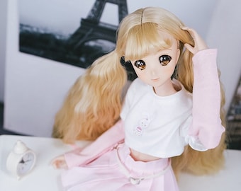 May include: A doll with long blonde hair styled in pigtails, wearing a white crop top with a cartoon bunny design, a pink pleated skirt, and white sneakers. The doll is sitting on a white shelf with a white clock in the background.