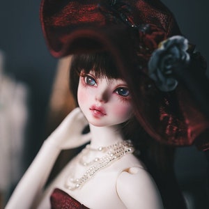May include: A white porcelain doll with blue eyes and dark hair wearing a red dress and a large red hat with a black rose.