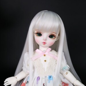 May include: A white doll with long, straight, white hair and green eyes. The doll is wearing a white dress with a pink bow and a lace trim. The dress has a colorful print of cartoon animals and stars.