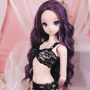 Puede incluir: Una muñeca de moda con largo cabello morado ondulado y ojos azul brillante. La muñeca lleva un conjunto de lencería de encaje negro. El fondo es rosa suave con acentos de tela blanca. La muñeca está arrodillada.