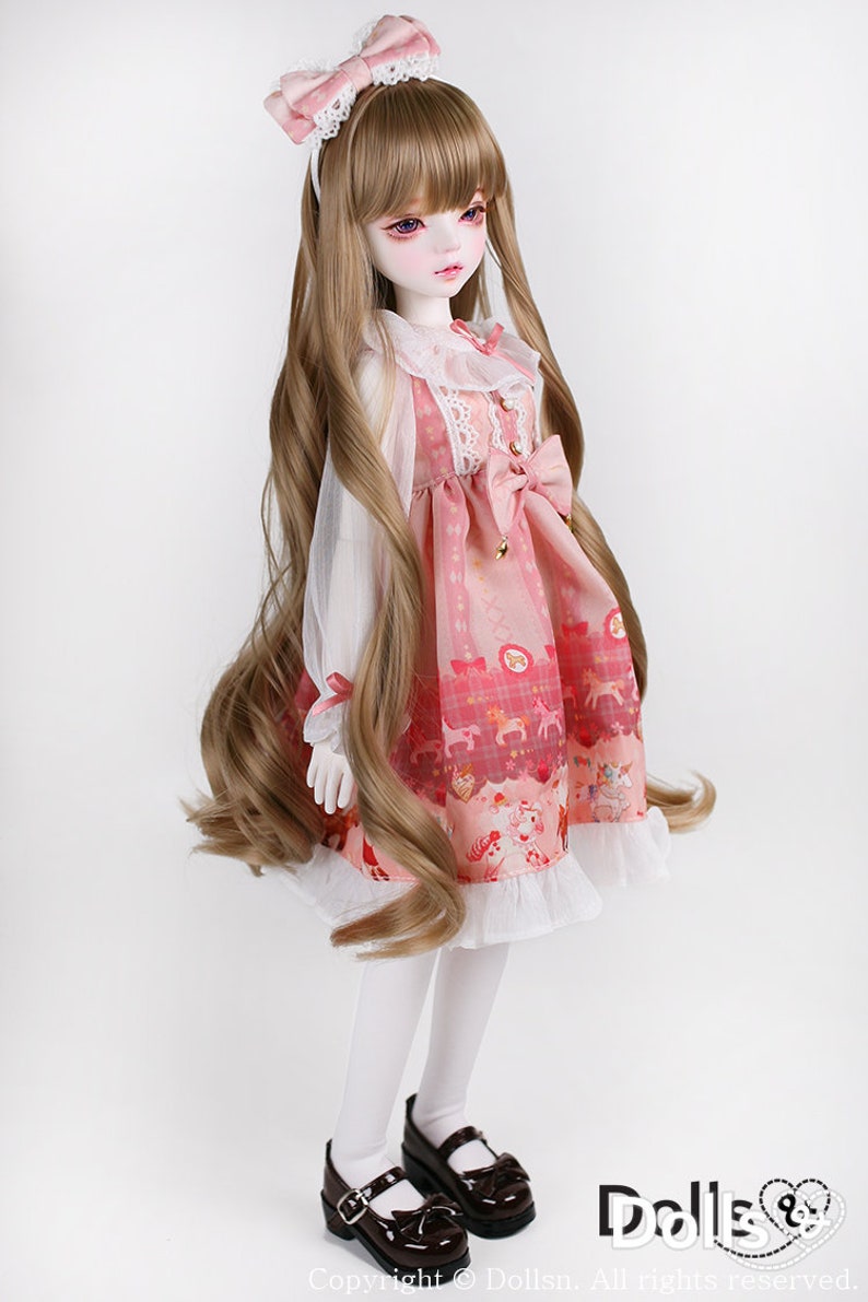 May include: A doll with long brown hair wearing a pink and white dress with a white lace collar and a pink bow. The doll is wearing white tights and black shoes.