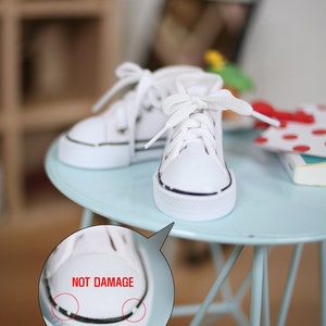 May include: A pair of white canvas high-top sneakers with black trim and white laces. The sneakers are sitting on a light blue table. The text "NOT DAMAGE" is written in red on a white circle.