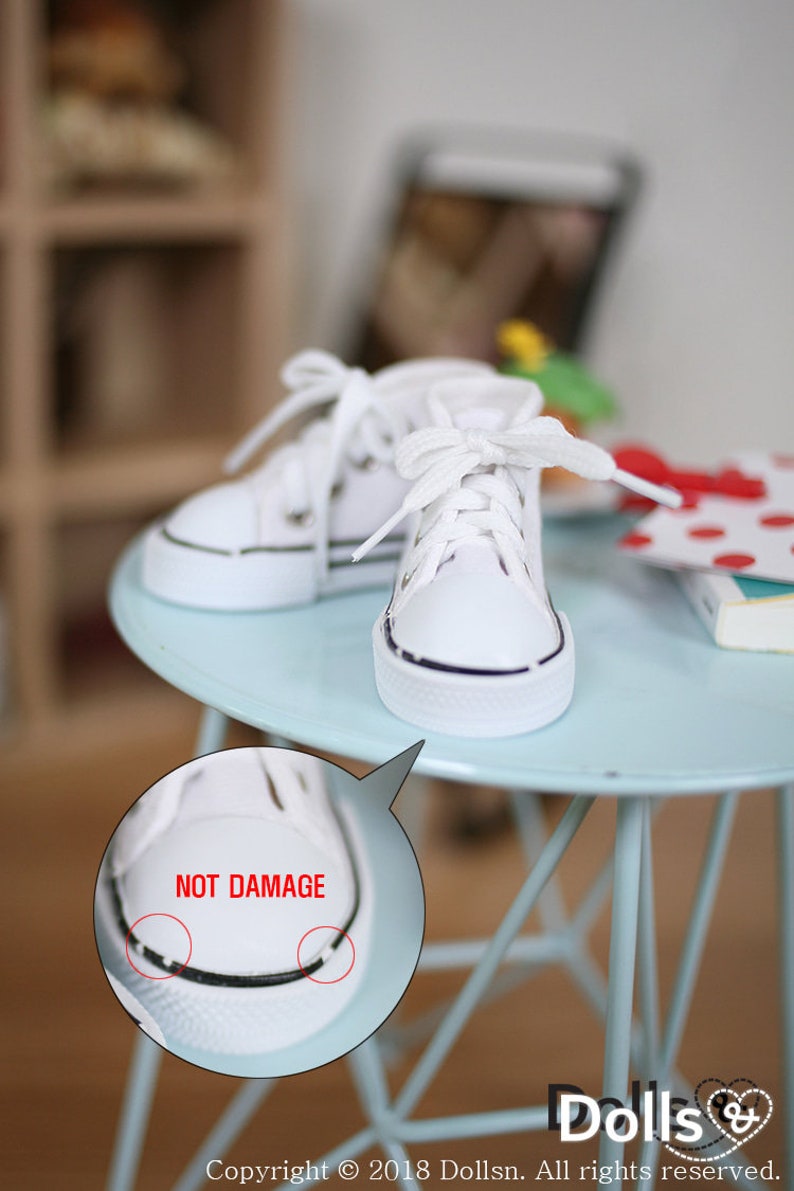 May include: A pair of white canvas high-top sneakers with black trim and white laces. The sneakers are sitting on a light blue table. The text "NOT DAMAGE" is written in red on a white circle.