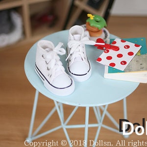 May include: A pair of white canvas high-top sneakers with black laces and a black trim around the sole, sitting on a light blue metal table. The table has a triangular base.