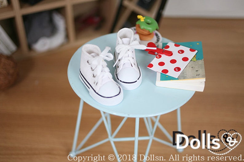 May include: A pair of white canvas high-top sneakers with black laces and a black trim around the sole, sitting on a light blue metal table. The table has a triangular base.
