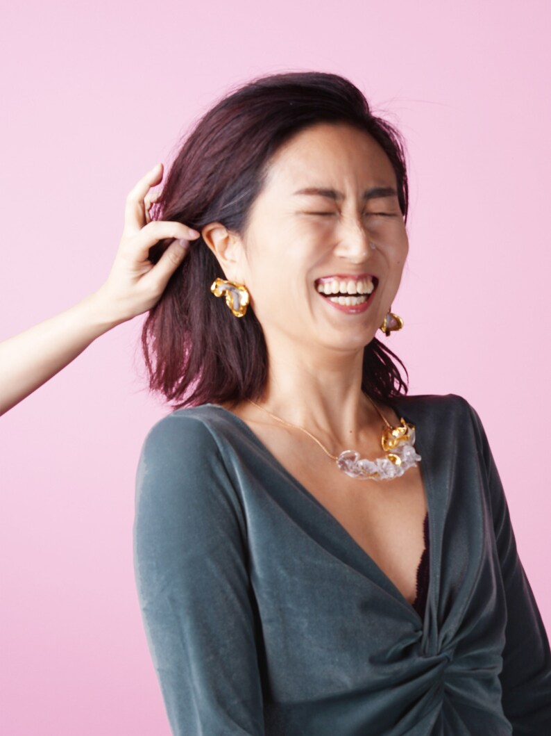 May include: A woman wearing a dark green velvet top with a clear and gold necklace and gold earrings. She is laughing and has her hair pulled back.