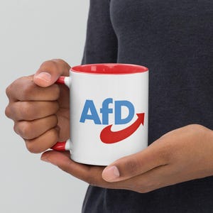 May include: White ceramic mug with a red rim. The mug has a blue and red logo that says "AfD" with a red arrow pointing to the right.