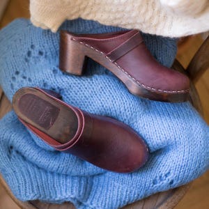 Puede incluir: Dos pares de zuecos de cuero burdeos con tacones de madera. Los zuecos están sobre una superficie de punto azul.