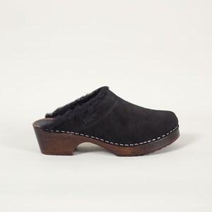 May include: Black suede clog with a shearling lining and a wooden sole. The shoe has a rounded toe and a low heel. The sole is dark brown with white stitching. The brand name is visible on the side of the shoe.