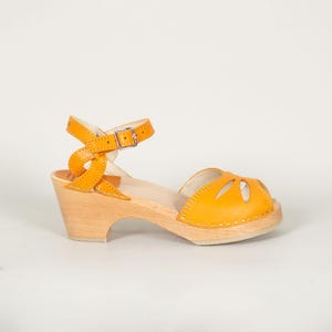 May include: A mustard yellow leather sandal with a peep-toe design, featuring a cutout pattern on the front and an adjustable ankle strap with a silver-toned metal buckle. The shoe is set on a light-colored wooden block heel and sole.