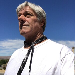 Puede incluir: Una persona con una camiseta blanca y un pa&ntilde;uelo negro con estampado de cachemira atado al cuello. El fondo es un cielo azul brillante con algunas nubes.