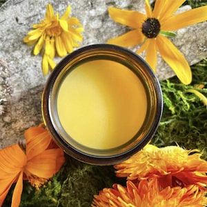 May include: A jar filled with a bright yellow substance, surrounded by yellow and orange flowers, green moss, and a gray stone. The jar's contents are smooth and opaque, contrasting with the natural textures around it. The scene evokes a sense of natural beauty.