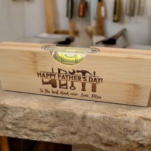 May include: A wooden level with a bubble vial, engraved with "HAPPY FATHER'S DAY!" and tools. The inscription reads "To the best dad ever. Love, Mia." The level is made of light-colored wood.