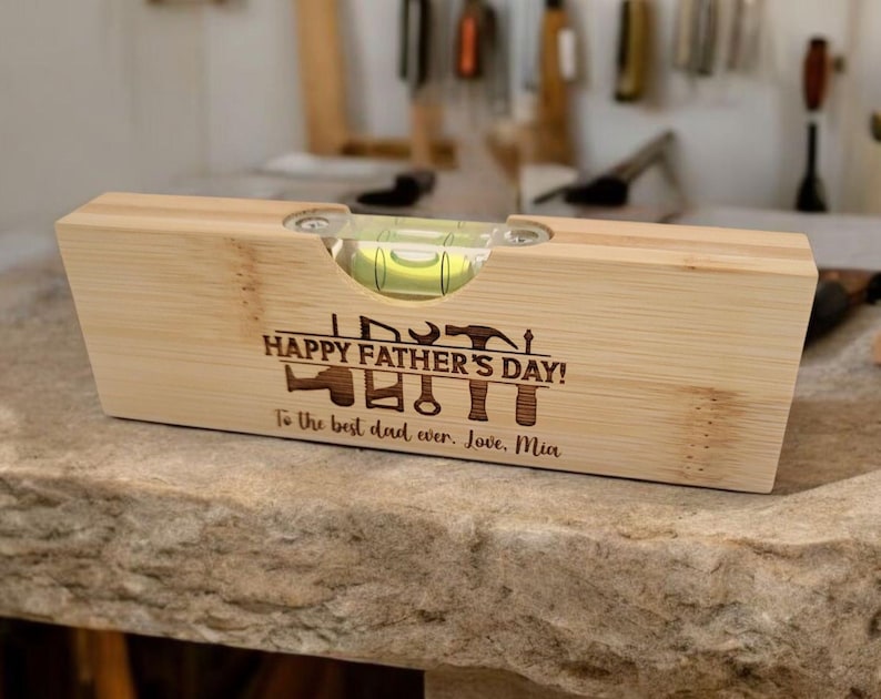 May include: A wooden level with a bubble vial, engraved with "HAPPY FATHER'S DAY!" and tools. The inscription reads "To the best dad ever. Love, Mia." The level is made of light-colored wood.
