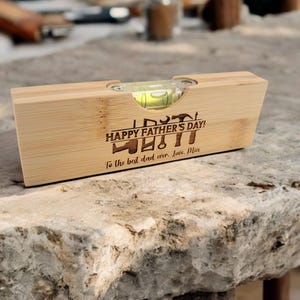 May include: A wooden level with a bubble vial, engraved with "HAPPY FATHER'S DAY!" and "To the best dad ever. Love, Mia." The level is made of light-colored wood and rests on a stone surface.