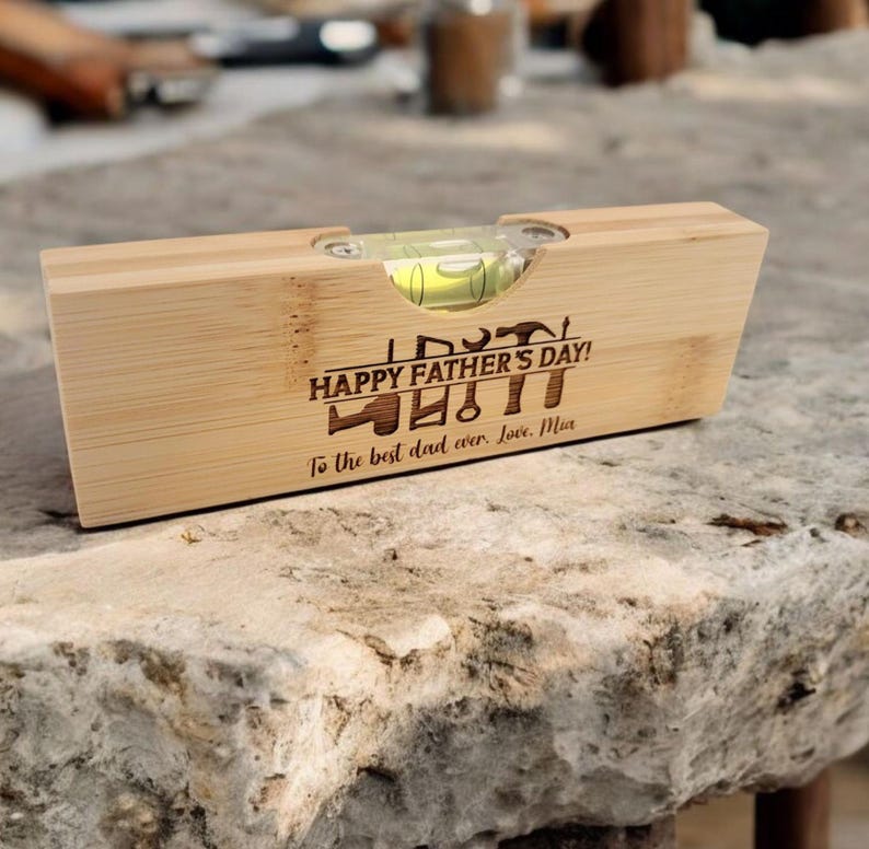 May include: A wooden level with a bubble vial, engraved with "HAPPY FATHER'S DAY!" and "To the best dad ever. Love, Mia." The level is made of light-colored wood and rests on a stone surface.