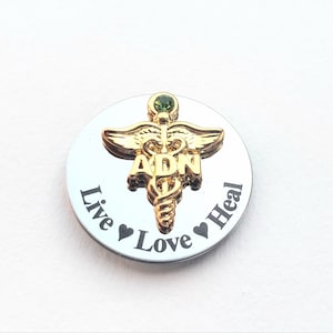 May include: A silver round pin with a gold medical caduceus symbol and a green gem. The pin says "Live Love Heal" and "ADN" in the center.