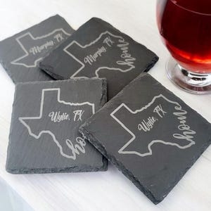 May include: Four square slate coasters with etched Texas state outlines and city names, plus the word "home". The dark grey coasters are displayed on a white surface. Each coaster measures approximately 10 cm x 10 cm.