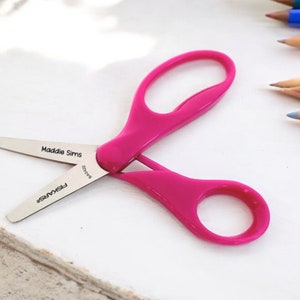 May include: Pink plastic children's scissors with a silver blade. The scissors are labeled "Maddie Sims" and "Fiskars".