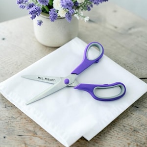 May include: A pair of scissors with purple handles and grey inner grips. The metal blade is engraved with the text Mrs. Milburn. The scissors are resting on a white folded cloth, with a potted plant visible in the background.