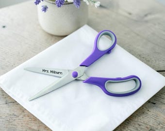 May include: A pair of scissors with purple handles and grey inner grips. The metal blade is engraved with the text Mrs. Milburn. The scissors are resting on a white folded cloth, with a potted plant visible in the background.