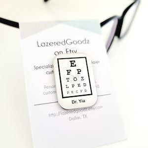 May include: A silver-colored eye chart pin with black lettering and a black border. The pin features the letters E, F, P, T, O, Z, L, P, E, D, and P, E, C, F, D. The text "Dr. Yiu" is below the chart.