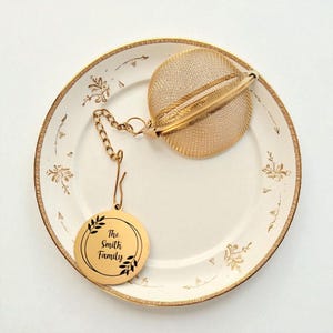 May include: A gold-colored tea infuser with a mesh ball and a chain, resting on a white plate with a gold floral pattern. A gold disc with the text "The Smith Family" is attached. The plate has a gold rim.