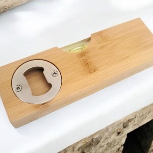 May include: A wooden bottle opener with a built-in level. The opener is made of light-colored wood and features a silver-colored bottle opener on one end. A small level is visible in the center. The item is on a white surface.