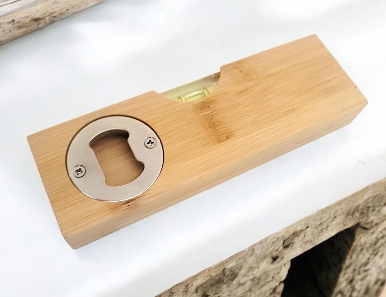 May include: A wooden bottle opener with a built-in level. The opener is made of light-colored wood and features a silver-colored bottle opener on one end. A small level is visible in the center. The item is on a white surface.