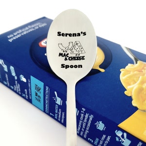 May include: A silver spoon with the text "Serena's Mac & Cheese Spoon" engraved on it. The spoon is resting on a blue and yellow box of macaroni and cheese.