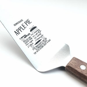 May include: A silver pie server with a wooden handle. The server has a recipe for homemade apple pie printed on the blade. The recipe includes 4 medium apples, 3/4 cup sugar, 1/4 cup flour, 1/2 tsp cinnamon, 1/4 tsp salt, and 1 tbsp lemon juice.