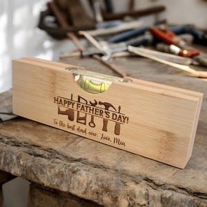 May include: A wooden level with a bubble vial, engraved with "HAPPY FATHER'S DAY!" and tools. The inscription reads "To the best dad ever. Love, Mia." The level is made of light-colored wood.