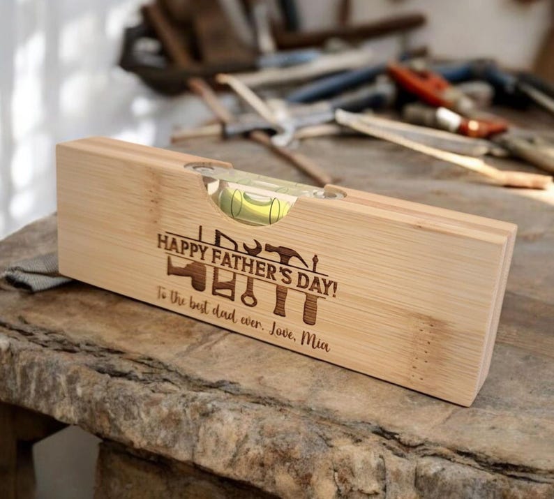 May include: A wooden level with a bubble vial, engraved with "HAPPY FATHER'S DAY!" and tools. The inscription reads "To the best dad ever. Love, Mia." The level is made of light-colored wood.