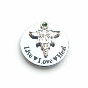 May include: Silver round charm with a green gem and a medical caduceus symbol. The charm has the words "Live Love Heal" engraved around the symbol.