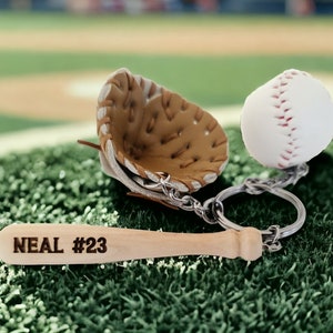 May include: A baseball-themed keychain with a brown leather baseball glove, a white baseball, and a wooden bat with the text "NEAL #23" engraved on it.