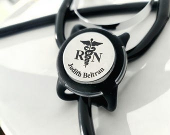 Puede incluir: Un estetoscopio negro con un dije plateado que dice "RN" y "Judith Beltran" con un símbolo de caduceo.