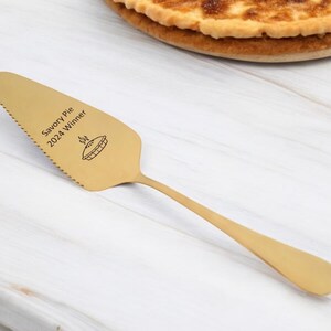 May include: A gold-colored pie server with a serrated edge and a long handle. The server has the text "Savory Pie 2024 Winner" and a pie graphic etched on the blade. A partially visible pie is in the background.