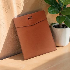 May include: A tan leather document sleeve with a pointed flap closure and the letters RM embossed in black on the front. The sleeve has a smooth finish and straight stitched edges, providing a structured appearance for holding papers or a tablet.
