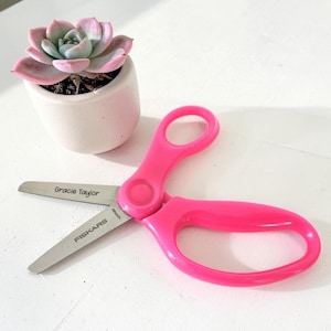 May include: Bright pink Fiskars scissors with a personalized name Gracie Taylor engraved on the metal blade. The scissors are positioned next to a small, light pink succulent plant in a matching matte ceramic pot, showcasing the tool's design.