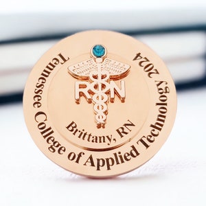 May include: A gold-colored round pin with a blue gemstone. The pin features a caduceus symbol with the letters "RN" and the text "Tennessee College of Applied Technology 2024 Brittany, RN".