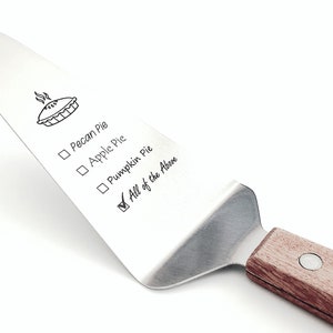 May include: A stainless steel pie server with a wooden handle. The server has a pie design and a checklist that says "Pecan Pie", "Apple Pie", "Pumpkin Pie", and "All of the Above" with a checkmark next to the last option.