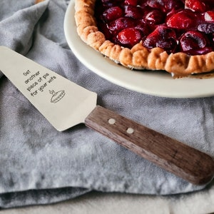 May include: A silver pie server with a wooden handle. The server has the text "Get another piece of pie for your wife" engraved on it.