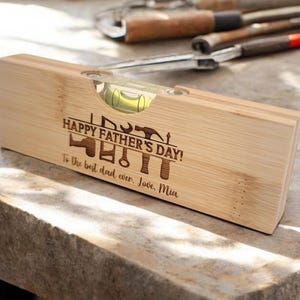 May include: A wooden level with a bubble vial, engraved with "HAPPY FATHER'S DAY!" and tools. The text "To the best dad ever. Love, Mia" is also engraved. The level is made of light-colored wood.