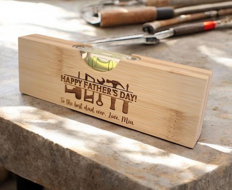 May include: A wooden level with a bubble vial, engraved with "HAPPY FATHER'S DAY!" and tools. The text "To the best dad ever. Love, Mia" is also engraved. The level is made of light-colored wood.